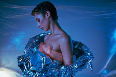 Metalik kumaşa bürünmüş bir kadın mavi bir arka plana karşı poz veriyor..