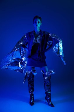 Mavi ışıklı stüdyodaki kadın geri dönüşümlü malzemelerden oluşan fütürist kıyafetler giyiyor.