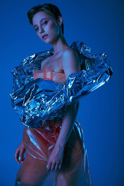Женщина позирует в синем свете, одетая в одежду из серебряной фольги и прозрачного пластика.