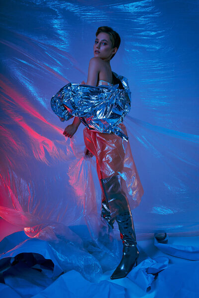 A woman in a silver jacket and black boots poses amidst a swirling sea of blue and red plastic.