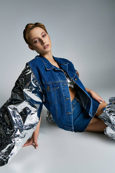 Женщина в джинсовой куртке с отражающими рукавами сидит на светло-сером фоне.