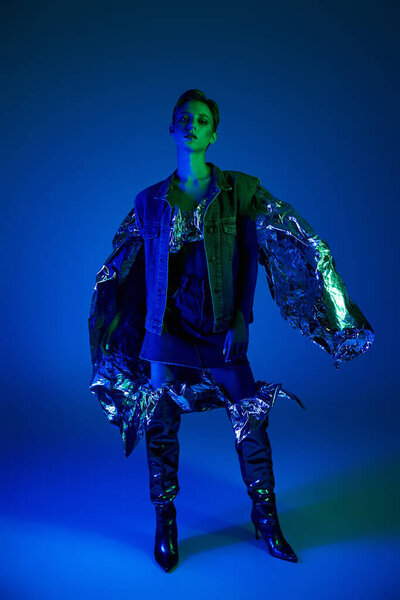 A woman in a denim jacket, standing in blue light, poses with foil wrapping her arms.