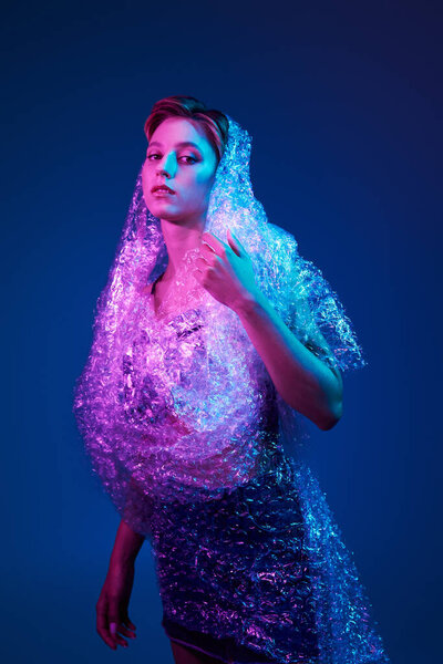 A woman enveloped in bubble wrap stands under vibrant neon lights.