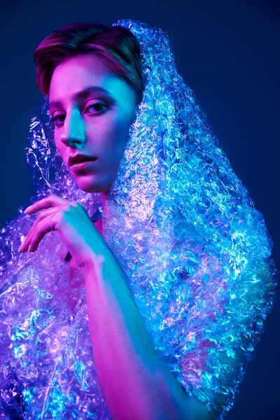 A woman in a studio setting is draped in bubble wrap, illuminated by vibrant pink and blue lights.