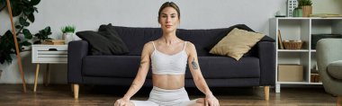 Modern bir dairede, etkin giyimli genç bir kadın yerde bağdaş kurup yoga yapıyor, sakin ve odaklanmış görünüyor.