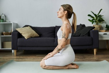 Aktif çalışan genç bir kadın, modern bir dairede yoga minderinde otururken meditasyon yapıyor..