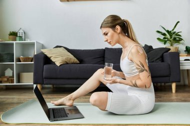 Vitiligo 'su olan genç bir kadın modern bir dairede paspasa oturmuş yoga yapıyor ve su içiyor..