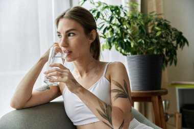 Vitiligo 'su olan genç bir kadın, aktif giyinmiş, modern bir dairede oturuyor ve yoga yaptıktan sonra su içiyor..