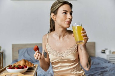 Saten bluzlu genç bir kadın yatağında otururken bir bardak meyve suyu ve çilek yiyor..