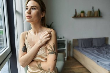 A young woman with vitiligo and tattoos stands by a window in her cozy bedroom, gazing out with a pensive expression. clipart