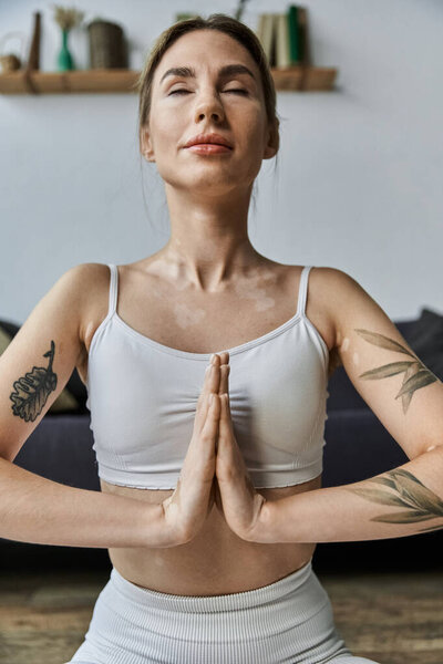 Молодая женщина в активной одежде сидит в современной квартире, практикуя йогу с закрытыми глазами, фокусируясь на медитации и психическом благополучии.