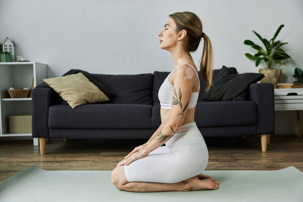 A young woman in active wear meditates while sitting on a yoga mat in a modern apartment.