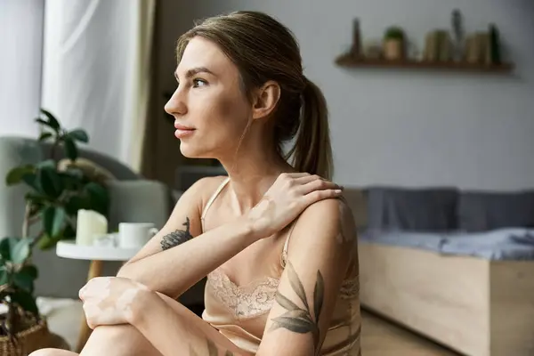 stock image A young woman with vitiligo and a tattoo sits in her bedroom, lost in thought.