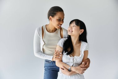 İki genç kadın, bir Afrikalı Amerikalı ve bir Asyalı, birbirlerine yakın dururlar, kollarını birbirlerine dolayıp, şefkatle sarılırlar..