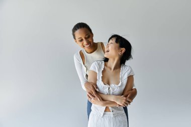 İki genç kadın, biri siyah, biri Asyalı, gri bir arkaplan önünde birbirlerine sarılırken gülüyorlar..