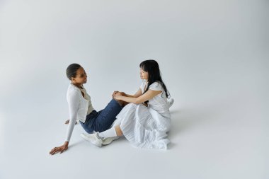 A young African American woman and an Asian woman share a tender moment while sitting on a white floor. clipart