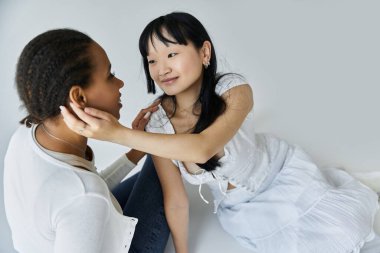 İki genç kadın, bir Afrikalı Amerikalı ve bir Asyalı birbirlerinin gözlerinin içine bakarken yüzleri birbirine yakın bir anı paylaşıyorlar..