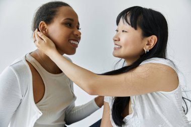 Siyah ve Asyalı bir kadın, gri bir arka planda, sevgi ve şefkat gösteren bir anı paylaşıyor.