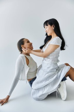 Genç bir Asyalı kadın, Afrikalı bir Amerikalı kadının kucağında oturuyor ve birbirlerine gülümsüyor..