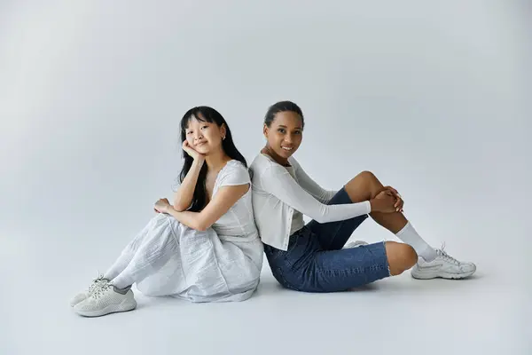 stock image A young Asian woman and a young Black woman sit back-to-back on a white background, smiling softly at the camera.