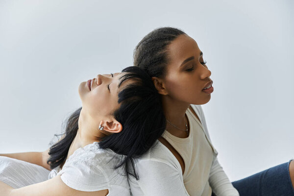 Две молодые женщины, лесбийская пара, сидят спина к спине с закрытыми глазами, разделяя нежный момент на сером фоне