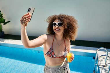 Bikinili bir kadın havuz kenarında selfie çekiyor, ferahlatıcı bir içecekle yaz gününün tadını çıkarıyor..