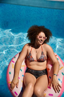 Güzel kıvırcık saçlı bir kadın havuzda gevşiyor, donut şeklinde bir şamandırada yüzüyor. Renkli bir bikini ve güneş gözlüğü takıyor..