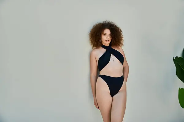 stock image A woman with curly hair, wearing a black swimsuit, poses confidently by a poolside wall on a summer day.