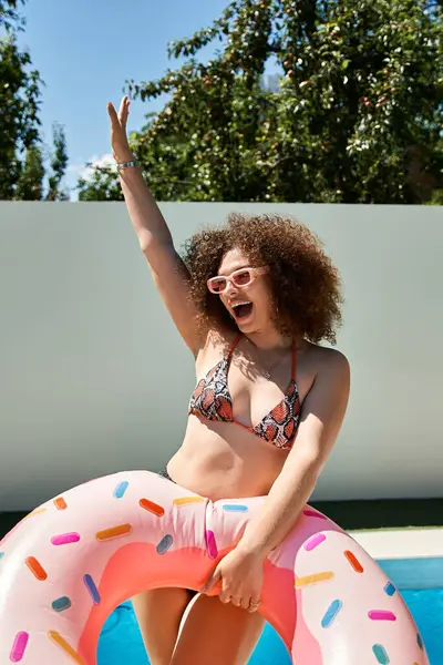 stock image A happy woman with curly hair, wearing a bikini and pink sunglasses, enjoys a donut float in the pool on a sunny day.