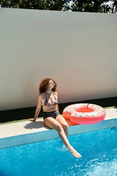 stock image A beautiful woman with curly hair relaxes on the edge of a pool, dangling her feet in the water, a pink donut float beside her.