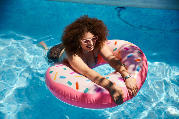 A woman with curly hair relaxes in a pool on a sunny day, enjoying the cool water and a colorful donut float.