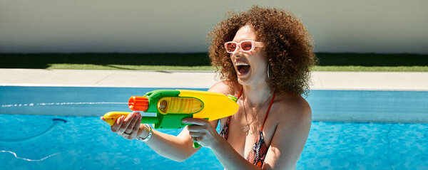 A woman with curly hair wearing a bikini enjoys a sunny day by the pool, playfully aiming a water gun.