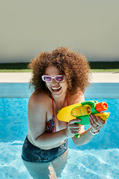 A woman in a bikini smiles as she holds a water gun in a pool.