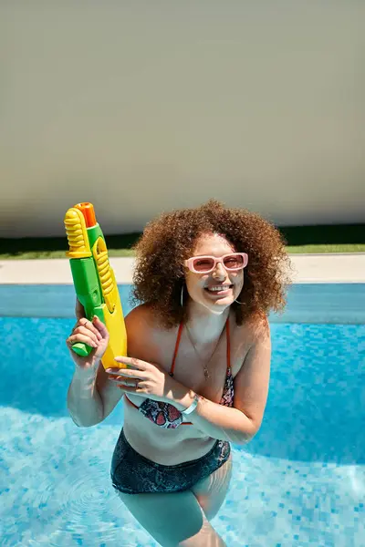 stock image A woman with curly hair and sunglasses smiles while holding a water gun in a pool.