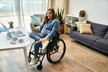 Tekerlekli sandalyedeki genç kadın gülümsüyor, modern dairede telefonla konuşuyor ve arka planda dizüstü bilgisayarı var.