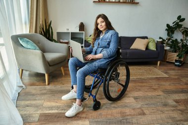 A young woman sits in her wheelchair, smiling, working on her laptop in a modern apartment. clipart