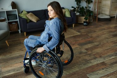 A young woman in a wheelchair sits in a modern apartment, looking thoughtfully towards the window. clipart