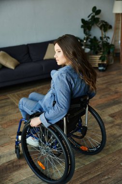 A young woman in a wheelchair sits in her modern apartment, looking thoughtfully toward the side. clipart