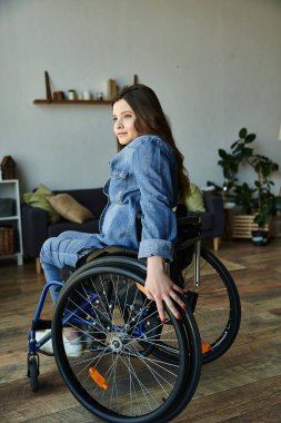Tekerlekli sandalyedeki genç bir kadın modern bir dairede oturuyor, düşünceli bir ifadeyle yan tarafa bakıyor..