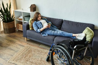 A young woman in a wheelchair lounges comfortably on a couch in her apartment, checking her phone. clipart