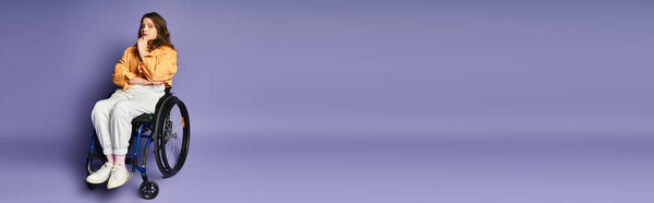 Молодая женщина в повседневной одежде задумчиво сидит в инвалидном кресле на фиолетовом фоне..