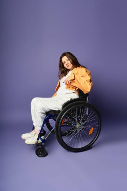 A young woman in a wheelchair and casual clothing poses in a studio with a purple background. clipart