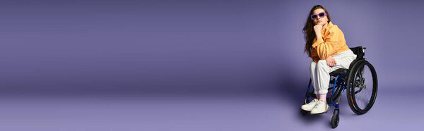 Молодая женщина в инвалидной коляске и в повседневной одежде позирует в студии с фиолетовым фоном.