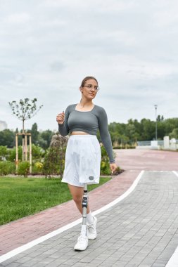 Protez bacaklı genç bir kadın açık havada spor yapmayı kucaklıyor ve manzaralı bir yolda rahatça egzersiz yapıyor..