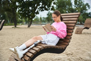 Spor kıyafetleri içinde genç bir kadın dışarıda rahatlıyor, eşsiz bir bankta oturmuş kitap okuyor..
