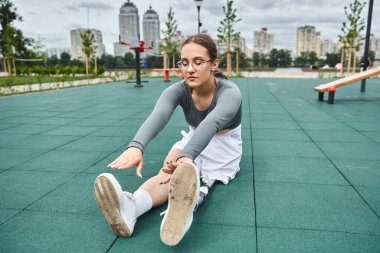 Genç bir kadın spor kıyafetleriyle spor yapıyor, bacaklarını şehir parkında yeşil bir spor salonunda açıyor..