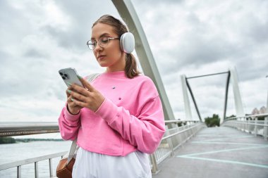 Telsiz kulaklıklı genç bir kadın köprüde akıllı telefon kullanıyor.