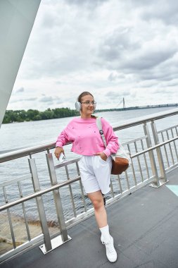 A young woman in sportswear listening music by the water, embracing her strength and vitality. clipart