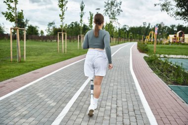 Protez bacaklı kararlı genç bir kadın dışarıda spor kıyafetleriyle egzersiz yapıyor, doğayı kucaklıyor..