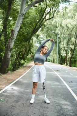 Genç bir kadın dışarıda güneşli bir günün tadını çıkarıyor. Spor kıyafetlerini protez bacaklarıyla esnetip egzersiz yapıyor..